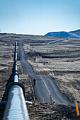 View of a long pipeline stretching across the stark, desolated landscape next to a narrow road under a clear sky, Nesjavellir, Grímsnes- og Grafningshreppur, Iceland.
