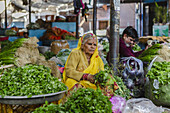 Bikaner, India - 24 June 2025: View of a woman in a yellow sari amidst a vibrant market, surrounded by fresh green produce and the bustling life of local vendors.