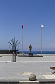 View of a bronze statue stands proudly on a stone plinth between two flags against the backdrop of a blue sky and sea, Girne, Northern Cyprus.