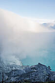 View of ethereal clouds swirling around rugged terrain, shrouding the turquoise lake in a mystical haze, a scene of raw natural beauty, Ijen Volcano Crater, East Java, Indonesia.