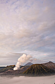 View of smoky tendrils rise from the volcanic peak, piercing the swirling clouds below, in a landscape of rugged beauty, Mount Bromo Volcano, East Java, Indonesia.