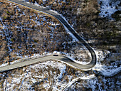 Luftaufnahme einer kurvenreichen Asphaltstraße, die sich durch eine Landschaft schlängelt, in der Flecken von schmelzendem Schnee mit dem kräftigen Braun und Grün der Bäume kontrastieren, mit einem einsamen Auto, das durch die Kurve fährt, Zagreb, Kroatien