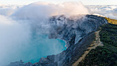Aerial view of ethereal clouds swirling around the rugged Ijen Volcano Crater, embracing its turquoise lake with a mystical allure, Ijen Volcano Crater, East Java, Indonesia.