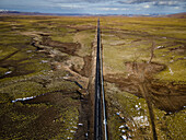 Luftaufnahme einer geraden, dunklen Straße, die durch eine lebendige, hügelige Landschaft mit erdigen Braun- und leuchtenden Grüntönen unter einem weiten Himmel führt, Mosfellsb?r, Island.