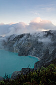 View of the stark, rocky landscape where wisps of clouds dance above a vibrant turquoise lake, a scene of otherworldly beauty, Ijen Volcano Crater, East Java, Indonesia.