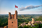 Aerial view of a medieval tower with a waving flag, surrounded by rolling hills dotted with terracotta roofs and lush green trees, San Miniato, Tuscany, Italy.