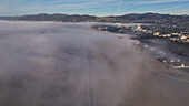 Aerial view of a thick fog blanketing the beach, with the city skyline barely visible in the distance, creating an ethereal coastal landscape, Los Angeles, California, United States.