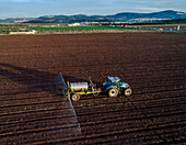 Luftaufnahme eines Traktors, der Wasser auf ein Feld sprüht, wobei die dunkle Erde mit dem hellen Himmel kontrastiert, Kabul, Norddistrikt, Israel.