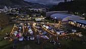 Aerial view of a bustling night market, illuminated by strings of lights, contrasting against the dark silhouettes of the surrounding hills, Nelson, Nelson Region, New Zealand.