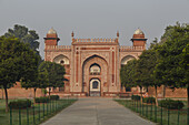 View of an ornate red sandstone structure framed by lush greenery along a pathway, evoking a sense of timeless beauty and historical grandeur, Agra, Uttar Pradesh, India.