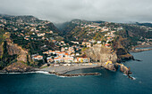 Luftaufnahme des Küstendorfs, das sich an die Klippen schmiegt, wo bunte Gebäude auf den dunkel schimmernden Ozean treffen, Ponta do Sol, Madeira, Portugal.