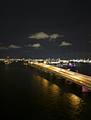Luftaufnahme einer Brücke, die sich über dunkle Gewässer unter einem wolkenverhangenen Nachthimmel erstreckt, mit schimmernden Lichtern entlang der Struktur und blinkenden Lichtern der Stadt in der Ferne, Miami, Florida, Vereinigte Staaten
