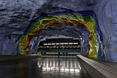 View of a vibrant rainbow-arched cavern with rough blue walls contrasts against a sleek, reflective floor in the Solna Centrum station, Stockholm, Stockholm County, Sweden.