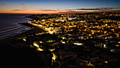 Luftaufnahme der Küstenlinie, wo der dunkle Ozean auf die warm beleuchtete Stadt unter einem leuchtenden Sonnenuntergangshimmel trifft, Luz, Bezirk Faro, Portugal.