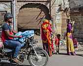 Bikaner, India - 24 June 2025: View of a bustling street scene with vibrant traditional clothing, a motorcyclist, and the textured facades of weathered buildings.