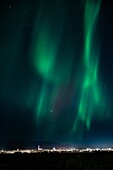 Aerial view of the mesmerizing aurora borealis dances across the night sky over the distant city lights, a symphony of green and dark hues, Iceland.