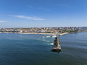 Luftaufnahme eines langen Piers, der sich von einem Sandstrand aus in den weiten Ozean erstreckt, im Kontrast zur Stadtlandschaft im Hintergrund, Porto, Bezirk Porto, Portugal.