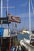 Girne, Cyprus - 25 June 2025: View of the Northern Cyprus flag fluttering proudly against the backdrop of Girne Castle, framed by the masts of boats in the harbor.