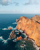 Luftaufnahme von zerklüfteten Klippen, die auf den unruhigen Ozean treffen, wo dunkle Felsen mit den warmen Farbtönen des Landes kontrastieren, Cani?al, Madeira, Portugal.