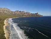 Luftaufnahme der Stelle, wo der Sandstrand auf den blauen Ozean trifft, unter dem wachsamen Blick der schroffen Berge, Kapstadt, Westkap, Südafrika.