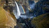 Aerial view of a cascading waterfall plunging into a rugged canyon, where a vibrant rainbow arcs across the misty spray near Haifoss, Iceland.