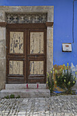 View of a weathered wooden door framed by stone, set against a striking blue wall, accented by vibrant yellow flowers and cobblestone street, Pano Lefkara, Larnaca, Cyprus.