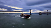 Hafen von Oakland, Vereinigte Staaten - 07. April 2025: Luftaufnahme eines kolossalen Frachtschiffs, das unter der majestätischen Bay Bridge durch den stählernen Ozean fährt.