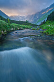 Blick auf einen ruhigen Fluss, der sich durch üppiges Grün zu hoch aufragenden Bergen unter einem wolkenverhangenen Himmel schlängelt, eine ruhige Flucht, Vysoké Tatry, Region Prešov, Slowakei.