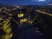Edinburgh, United Kingdom - 16 June 2025: Aerial View of Edinburgh Castle illuminated at night, where vibrant yellow spotlights cut through the dark, contrasting with the muted city lights, under a twilight sky.