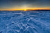 View of a vast, windswept expanse of blue-tinted snow drifts sculpted by the elements under the warm glow of the rising sun, Finnmark, Norway.