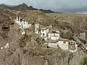 Aerial view of the stark white structures of the Chemrey Monastery cling to the rugged, ochre-colored cliffs under a cloudy sky, Leh, Ladakh, India.