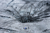 Aerial view of a massive glacial sinkhole with dark textures and four people standing near the edge, Selfoss, Sveitarfélagið Árborg, Iceland.