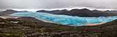 View of a vibrant turquoise glacier snakes between dark, rugged mountains under a brooding sky, surrounded by stark, volcanic terrain, Iceland.