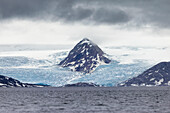 Blick auf einen spitzen Berg, der sich über einen riesigen Gletscher erhebt, wo das Eis auf das dunkle, gekräuselte Meer unter einem schweren, bedeckten Himmel trifft, Svalbard und Jan Mayen.