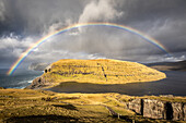 View of a vibrant rainbow arcs over a rugged island with steep cliffs and golden grass contrasting with the deep blue sea, Vágar, Faroe Islands.