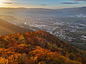 Luftaufnahme der feurigen Farben des Herbstes, die die Landschaft einhüllen, ein Wandteppich aus Orange und Gold, der sich auf die im Tal gelegene Stadt zubewegt, Bansk? Bystrica, Region Bansk? Bystrica, Slowakei.