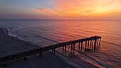 Luftaufnahme des Piers von Saint Johns County, der sich unter einem leuchtenden, in Orange-, Rosa- und Violetttönen gehaltenen Himmel in den Ozean erstreckt, Saint Augustine Beach, Florida, Vereinigte Staaten.
