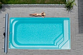Aerial view of a sunbather relaxing by the pool's turquoise waters, contrasted by the cool grey tiles and verdant lawn, creating a serene oasis, Strasbourg, Grand Est, France.