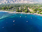 Aerial view of boats dotting the turquoise sea meeting the dark sandy beach alongside the verdant jungle under the distant mountains, Pemuteran, Bali, Indonesia.