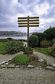 View of a weathered wooden signpost stands amidst a vibrant garden path, pointing towards unseen destinations against the backdrop of Fowey's coastal charm, Fowey, England, United Kingdom.