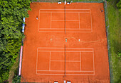 Luftaufnahme von zwei leuchtend orange-roten Tennisplätzen inmitten üppiger grüner Bäume, die einen auffälligen Kontrast von Farben und Texturen bilden, Ebersberger Forst, Bayern, Deutschland.