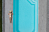 Aerial view of a woman in a swimsuit and hat laying beside a bright blue swimming pool, Strasbourg, Grand Est, France.