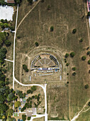 Aerial view of the ancient Roman Theatre, its weathered stone contrasting with the sun-baked grass and winding paths, Gubbio, Perugia Province, Italy.