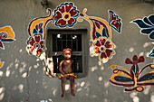 Chapai Nawabganj, Bangladesch - 14. Juni 2019: Blick auf ein Kind, das auf einem Fensterbrett sitzt, das mit lebhaften, handgemalten Blumenmotiven vor einer strukturierten Wand geschmückt ist.