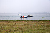 Bahmanbaria, Bangladesch - 30. Januar 2024: Blick auf kleine Boote, die über das ruhige, spiegelnde Wasser unter einem blassen, nebligen Himmel gleiten, eine ruhige Szene mit einer Mischung aus erdigen Braun- und gedämpften Blautönen.