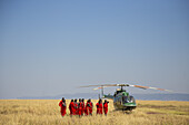 Narok, Kenia - 08. November 2018: Blick auf einen Hubschrauber, der auf der goldenen Savanne ruht, während die in leuchtendes Rot gehüllten Maasai-Krieger in starkem Kontrast zum ruhigen blauen Himmel stehen.