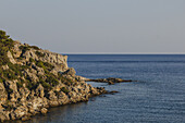 Blick auf zerklüftete Klippen und das ruhige azurblaue Meer unter klarem Himmel, eine zeitlose Szene natürlicher Schönheit und küstennaher Gelassenheit, Kallithea, Rodhes, Griechenland.