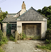 Lostwithiel, United Kingdom - 31 May 2025: View of a quaint, weathered building with a faded pink facade, a dark wooden garage door, and a crumbling chimney against a backdrop of lush greenery.