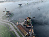 Aerial view of historic windmills rising through the early morning fog, creating an ethereal landscape of tradition and tranquility, Schansend, Noord-Holland, Netherlands.