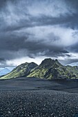View of stark black volcanic terrain stretches toward verdant mountains beneath a brooding sky, a landscape of raw beauty and dramatic contrasts, Kirkjubaejarklaustur, Skaftárhreppur, Iceland.
